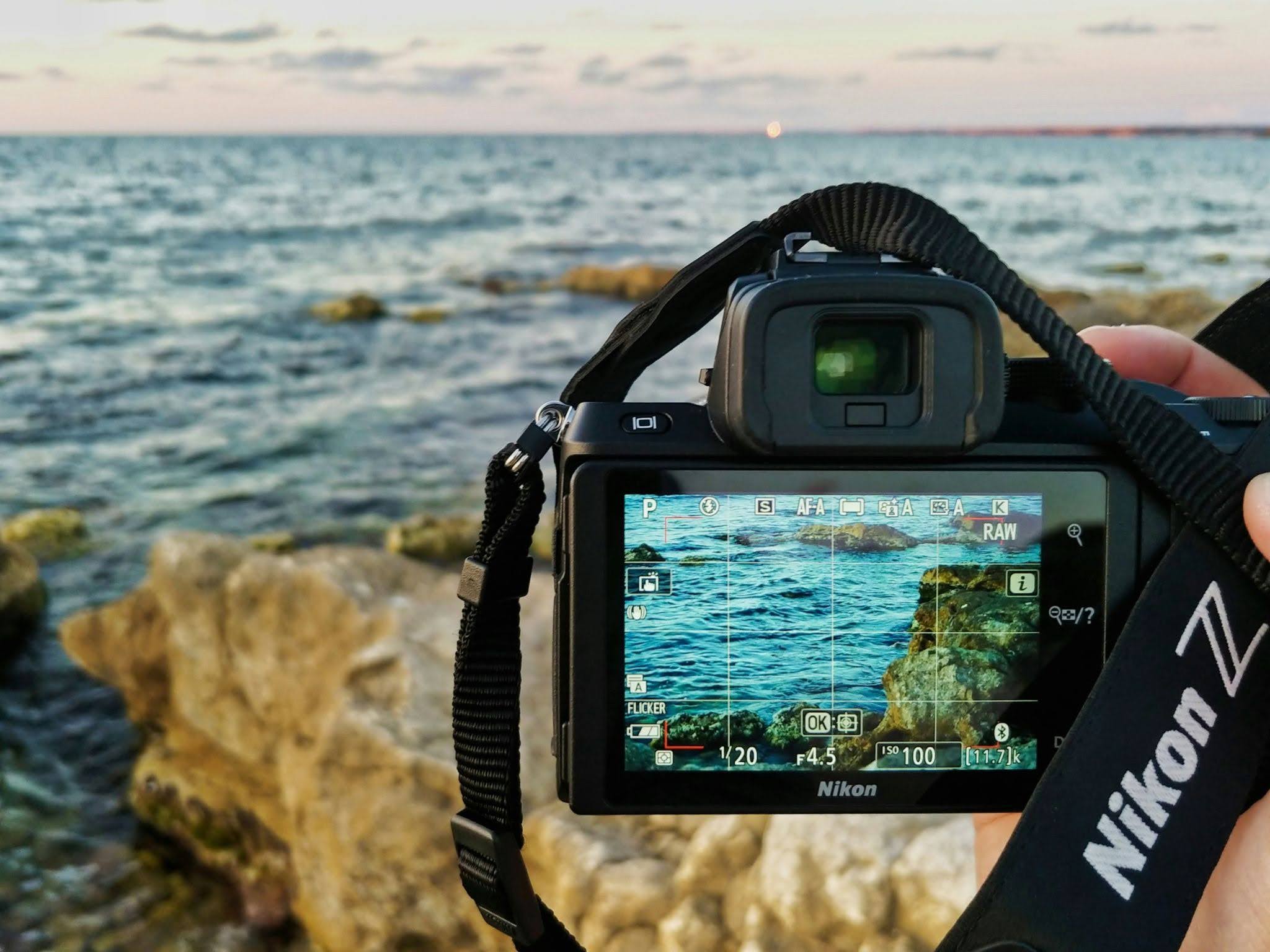Digital display of a Nikon camera showing an authentic sunset over a rocky coastline, emphasizing the beauty of untouched nature and low-impact exploration.